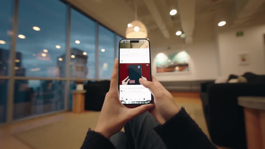 A first-person view of hands scrolling through a social media feed on a smartphone inside a modern room with large windows