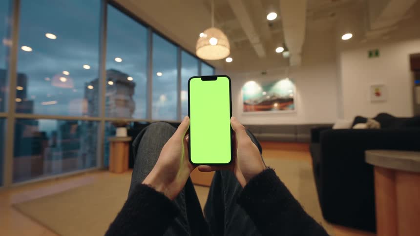 POV shot of a person holding a smartphone with a green screen in a modern lounge overlooking the city at dusk