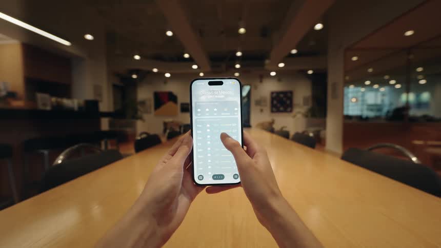 First-person point-of-view of hands holding a smartphone and scrolling through a weather forecast application in a modern office