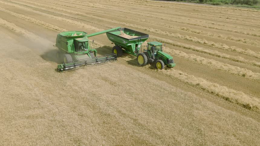 Aerial view of a combine harvester unloading freshly harvested grain into a tractor-pulled grain cart while working in a vast field