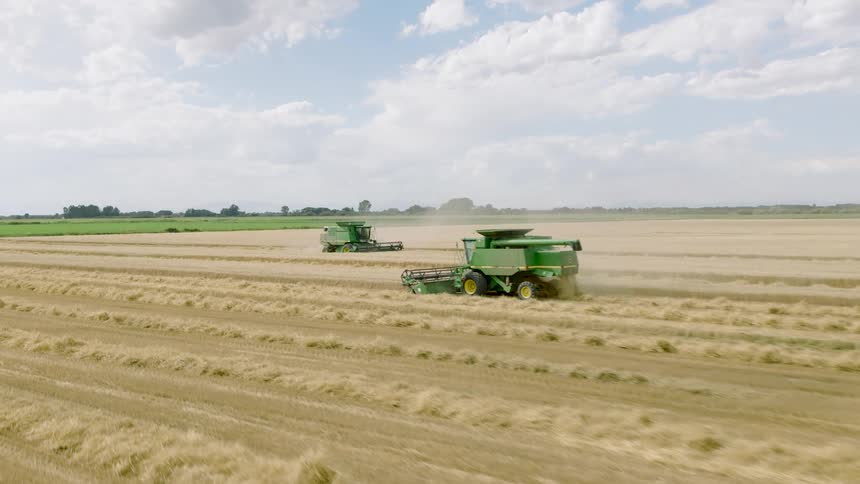 An aerial view shows two combine harvesters moving through a vast golden field, efficiently harvesting grain under a partly cloudy sky