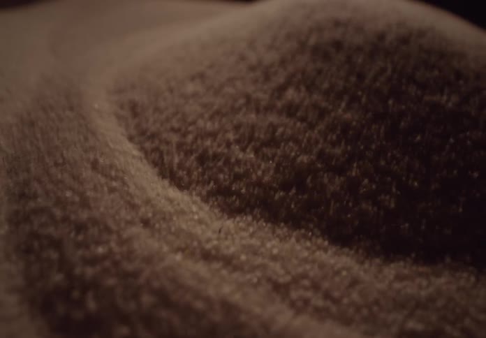 An abstract macro shot shows a gentle breeze blowing tiny grains of sand across a miniature dune in slow motion
