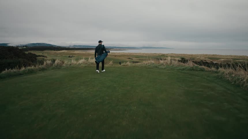 A golfer walks with his bag over a hill on a coastal links course, revealing the stunning landscape in an epic aerial shot