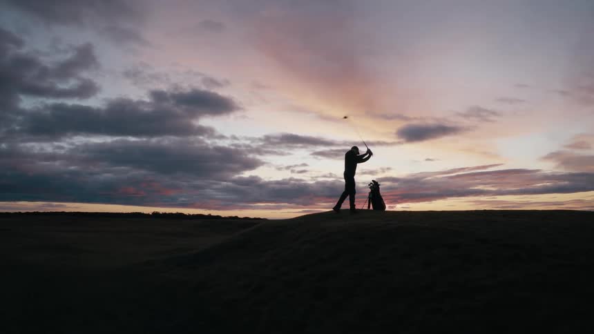 A silhouetted golfer takes a powerful swing in slow motion on a beautiful, expansive course against a dramatic sunset sky