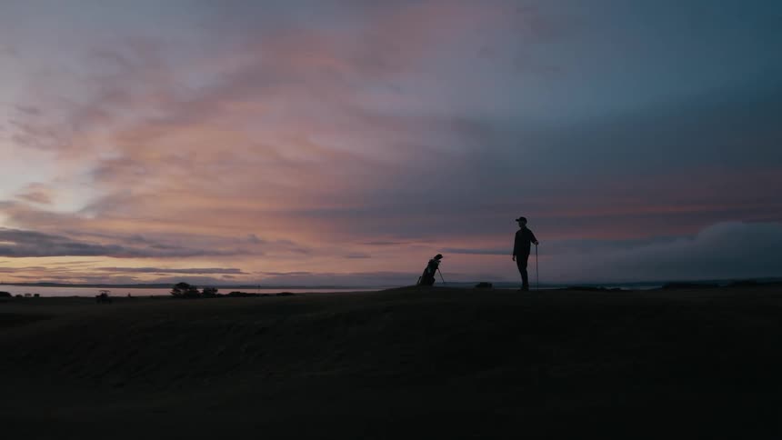 A silhouetted golfer walks onto a hill on a coastal golf course to admire the beautiful, dramatic sunset over the ocean