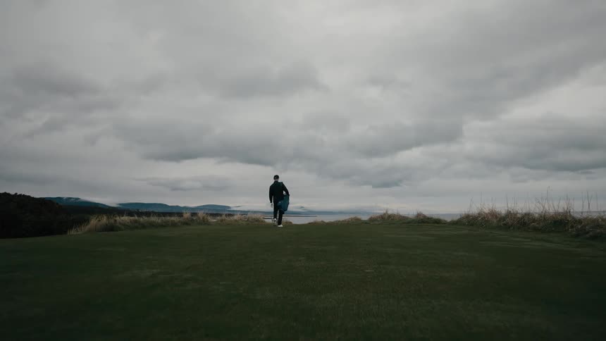 A lone golfer walks to the edge of a tee box, and the camera reveals a stunning coastal golf course under an overcast sky