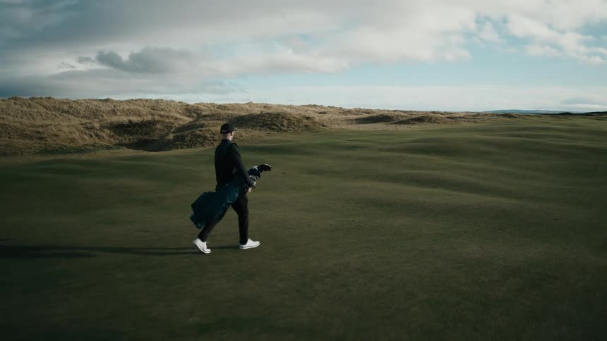 An aerial shot follows a lone golfer carrying his bag across a vast, scenic links course before revealing the beautiful coastline