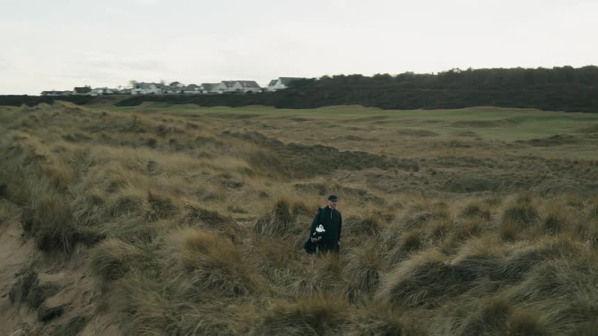 A lone golfer carries his clubs and walks in slow motion through the tall grass on the dunes of a seaside links course