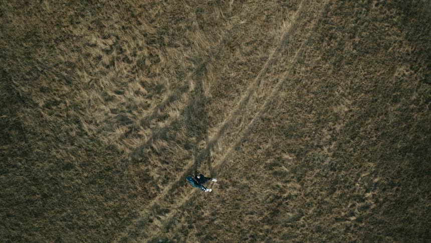 An aerial top-down view follows a lone snowboarder walking across a vast, dry grassy field