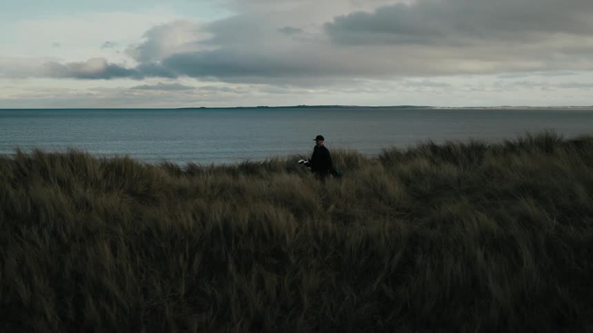 A lone golfer carries his clubs in slow motion as he walks across a windy, grassy dune overlooking the ocean