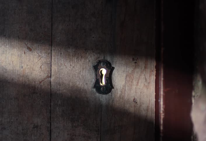 A single beam of sunlight illuminates a vintage keyhole on a weathered wooden door, with a mysterious light flickering through it