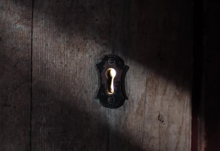 A flickering candle flame is visible through the rusty keyhole of a weathered wooden door