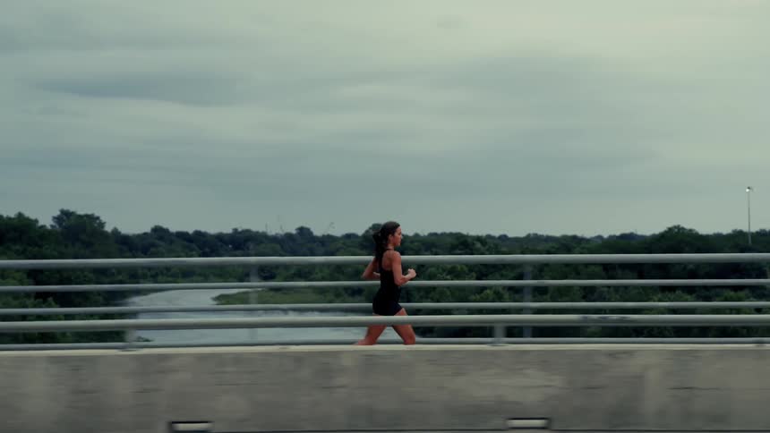 Slow motion side profile of a determined female athlete jogging across a bridge over a river on a cloudy day