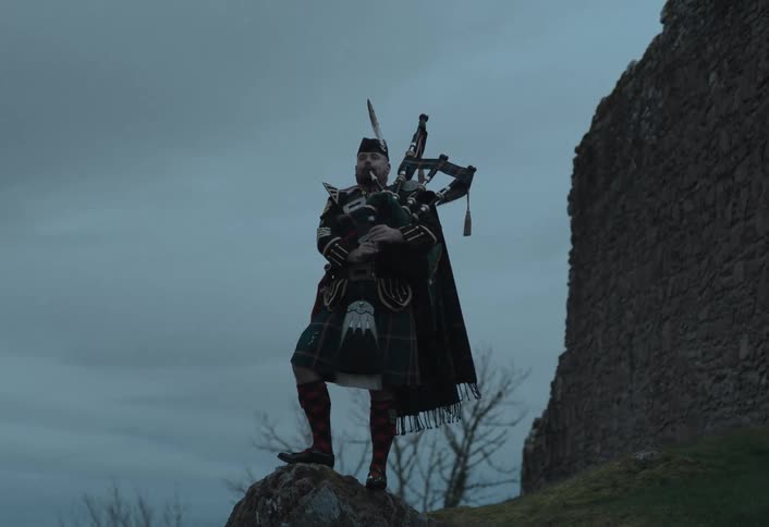 A traditional Scottish bagpiper plays a solemn tune on a rocky hill next to the ruins of a castle