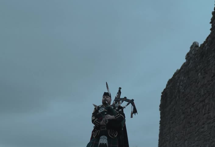 A low-angle shot captures a Scottish piper in a traditional kilt playing his bagpipes next to an ancient stone wall under an overcast sky