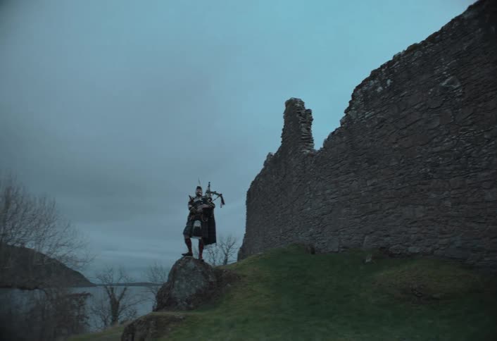A tracking shot reveals a Scottish bagpiper in a kilt standing stoically beside ancient castle ruins overlooking a loch