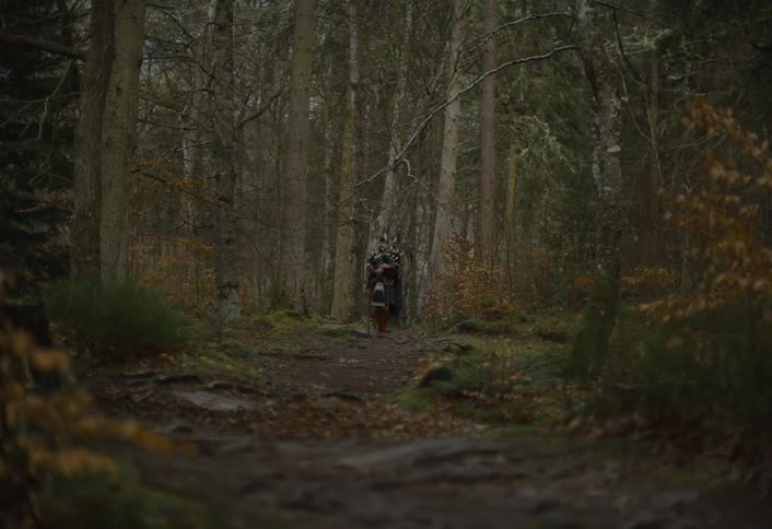 A lone Scottish bagpiper in a traditional kilt stands solemnly on a path in a dense, dark forest