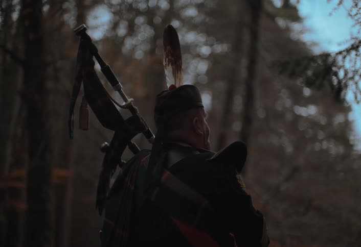 A Scottish bagpiper dressed in a traditional kilt and feathered cap stands solemnly in a dark forest