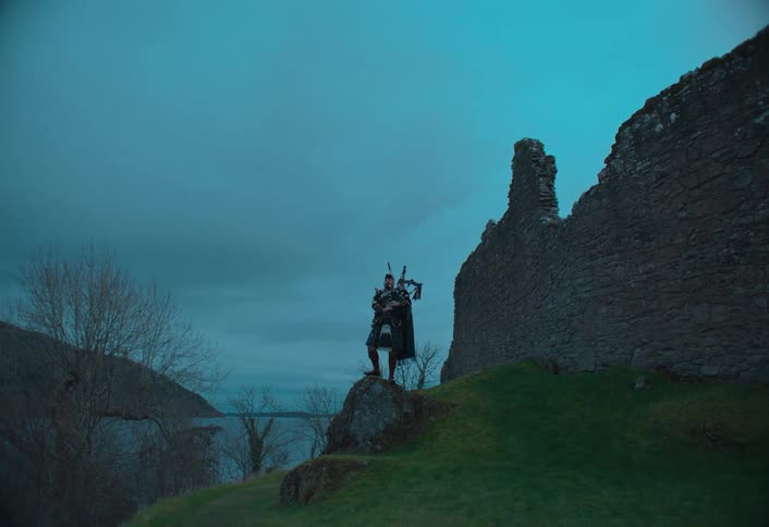 A lone Scottish bagpiper in a traditional kilt plays his instrument atop a hill next to ancient castle ruins overlooking a loch at dusk
