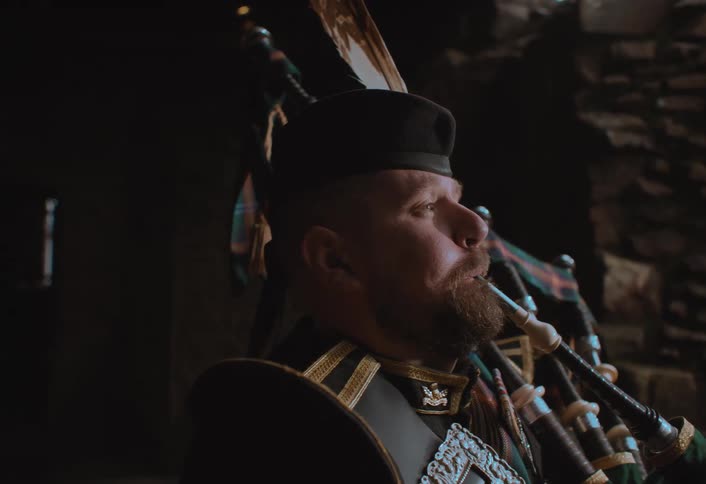 A solemn Scottish man in traditional uniform plays the bagpipes in a dark, atmospheric stone hall