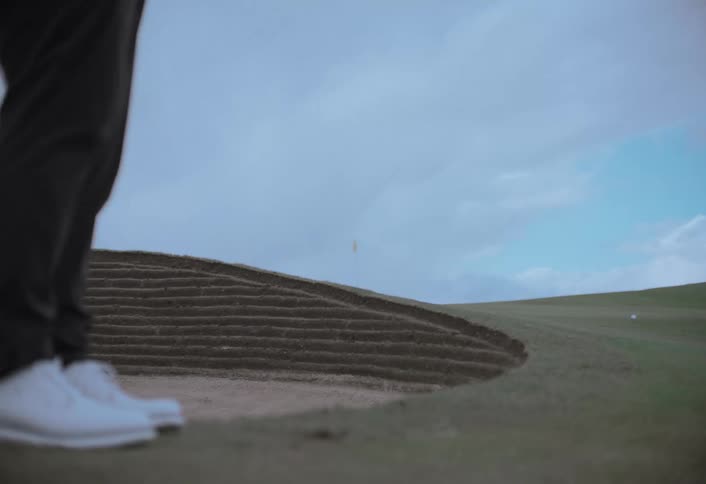 A professional golfer chips a golf ball onto the green in slow motion next to a large sand bunker