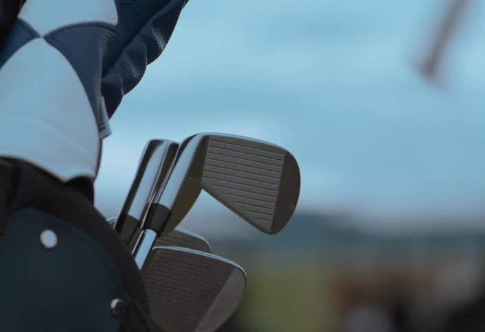 A golfer selects an iron from their golf bag with the course and carts visible in the background