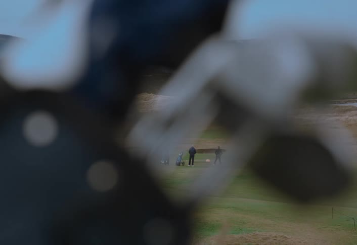 A distant shot of men playing golf on a green course, framed by out-of-focus golf clubs in the foreground as one man takes a swing in slow motion
