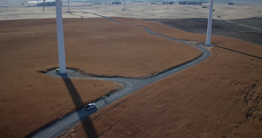 High aerial view of a wind farm in a golden field, showing a winding road where two workers stand beside a truck at the base of a turbine