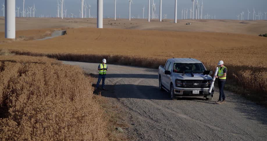 Rising aerial shot of two engineers reviewing blueprints on a truck amidst a sprawling wind turbine farm