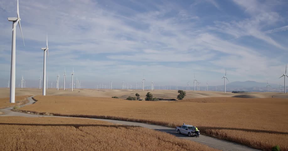 A pair of energy workers stand by their truck inspecting a sprawling wind farm on golden rolling hills under a partly cloudy sky