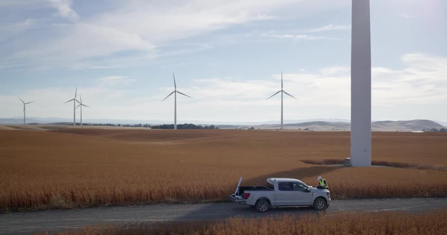 An engineer reviews plans on the hood of his truck at a large wind farm in a sunny, rural field as the camera pulls back