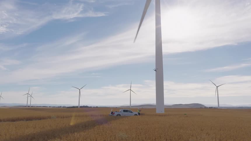 A technician uses a drone to perform a maintenance inspection on a giant wind turbine in a sunny, golden field at a wind farm