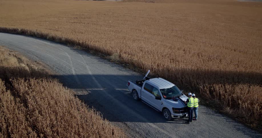 Aerial view of two engineers in hard hats reviewing blueprints on their truck at a large wind farm in a golden field