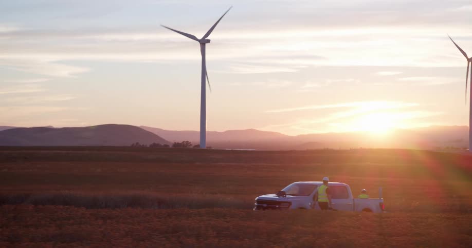 Wind turbine technicians stand by their truck in a vast golden field with windmills spinning during a beautiful sunset