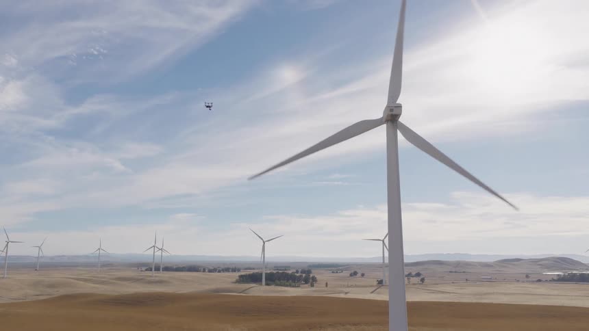 A drone hovers near a large spinning wind turbine at a wind farm under a sunny sky