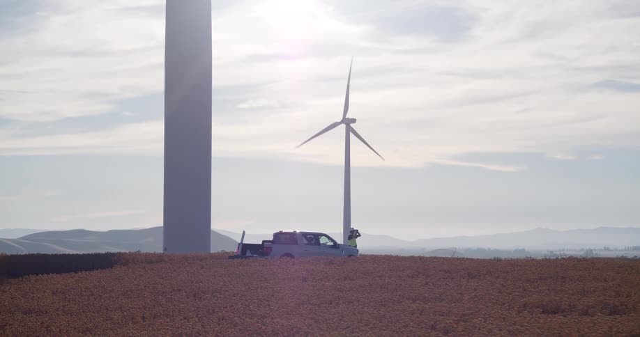 Two wind turbine engineers stand by their truck at the base of a large turbine in a golden field on a sunny day