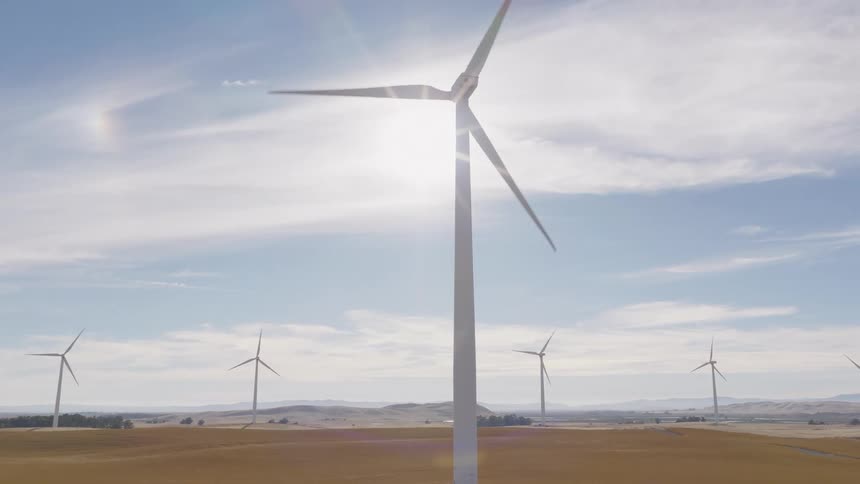 Sun shines brightly behind a slowly spinning wind turbine on a vast wind farm in a golden field on a clear day