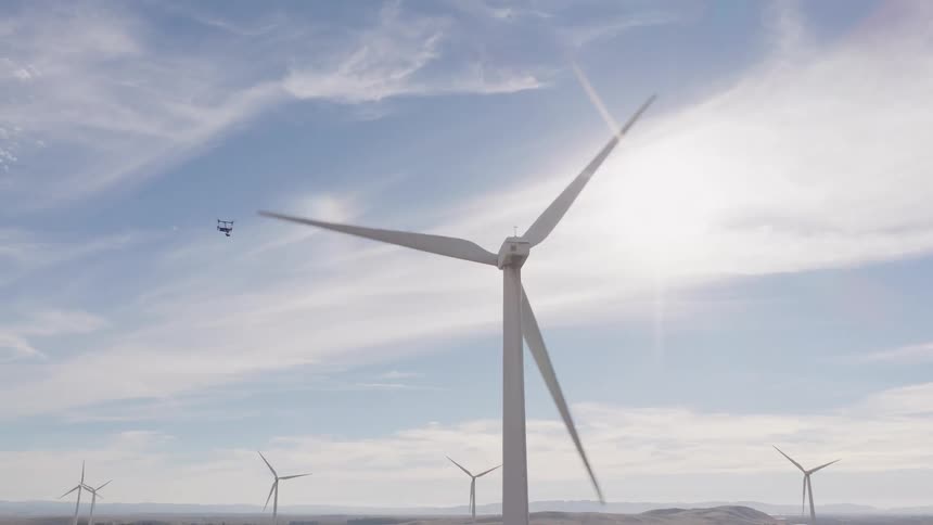 An inspection drone flies near a massive wind turbine with the sun flaring brightly behind it on a sprawling wind farm