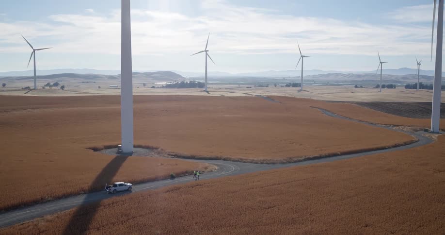 Aerial view of a cyclist riding down a winding rural road through a golden field dotted with large, spinning wind turbines