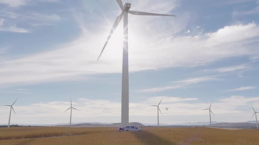 Massive wind turbines generate clean energy in a golden field as a drone performs an inspection on a sunny day