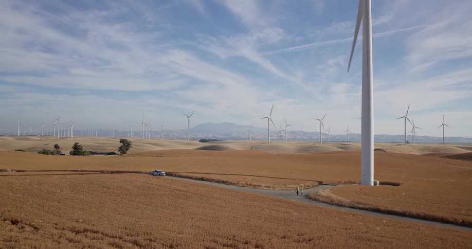 A sprawling wind farm with dozens of spinning turbines dominates the golden, rolling hills as two workers walk on a dirt road below