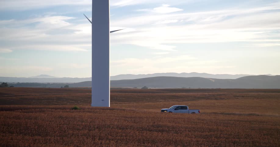 A white pickup truck is parked in a golden field at the base of a slowly spinning wind turbine with rolling hills in the background