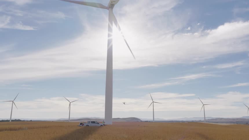 Engineers launch a drone to perform a maintenance inspection on a massive wind turbine at a wind farm in a sunny field