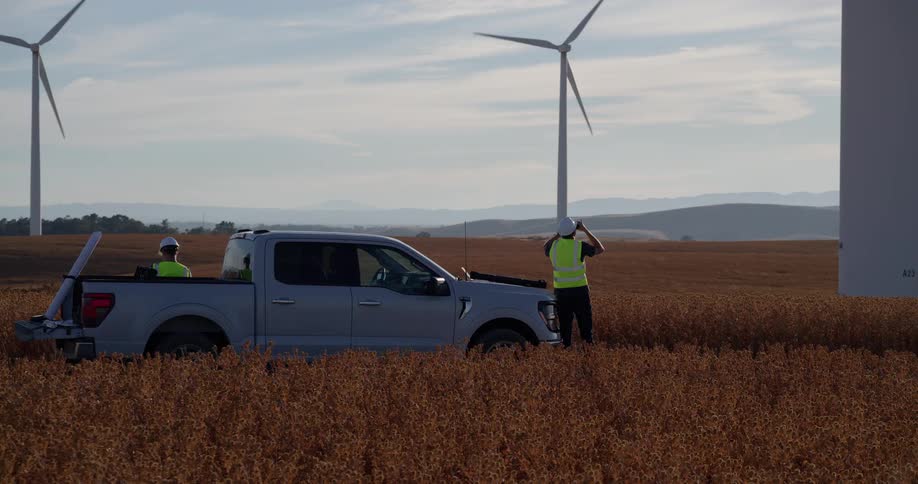 A team of engineers in safety vests survey a large wind farm from their pickup truck in a vast rural field