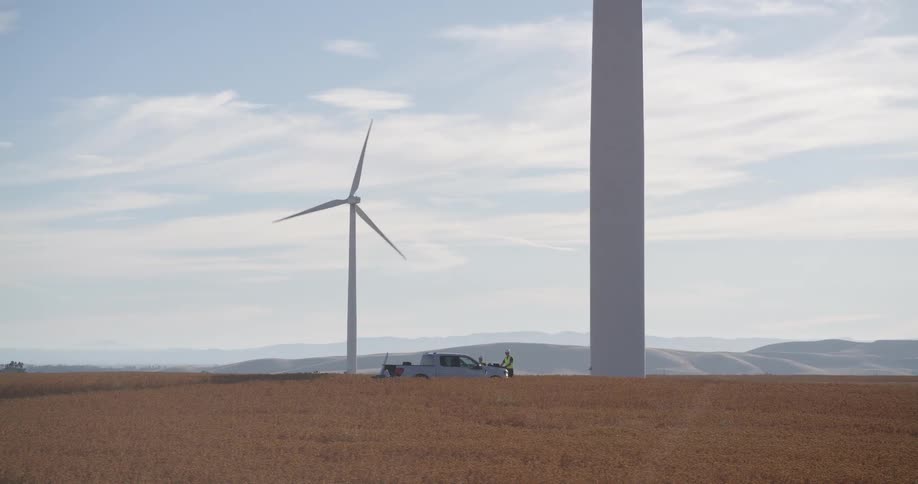 Wind turbine technicians with their truck inspect a massive windmill on a sprawling wind farm in a golden field