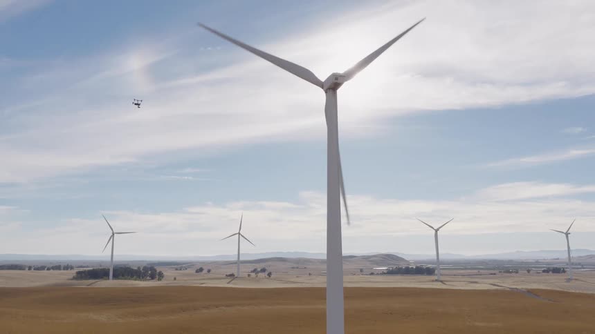 A drone hovers near a large wind turbine as its blades spin in the bright sunlight on a vast wind farm in a rural landscape