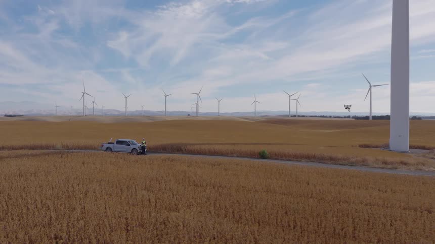 A drone flies up to inspect a large wind turbine on a wind farm as a technician watches from below