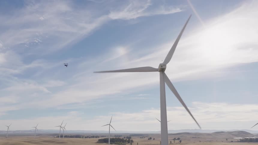 A drone flies away from a spinning wind turbine at a large wind farm across a sunny, dry, rolling landscape