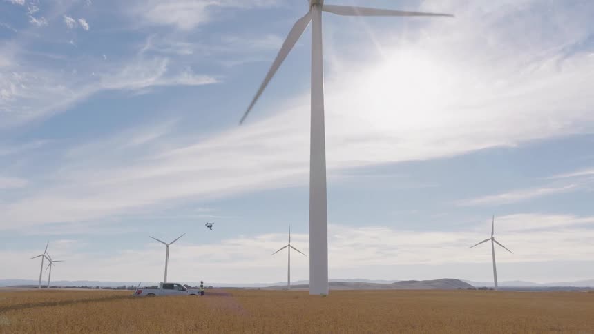 A drone hovers near a giant spinning wind turbine in a sunny field during an industrial inspection at a renewable energy farm