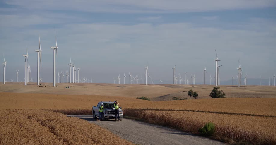 Two wind turbine technicians in safety gear collaborate over blueprints on their truck amidst a vast, rural wind farm